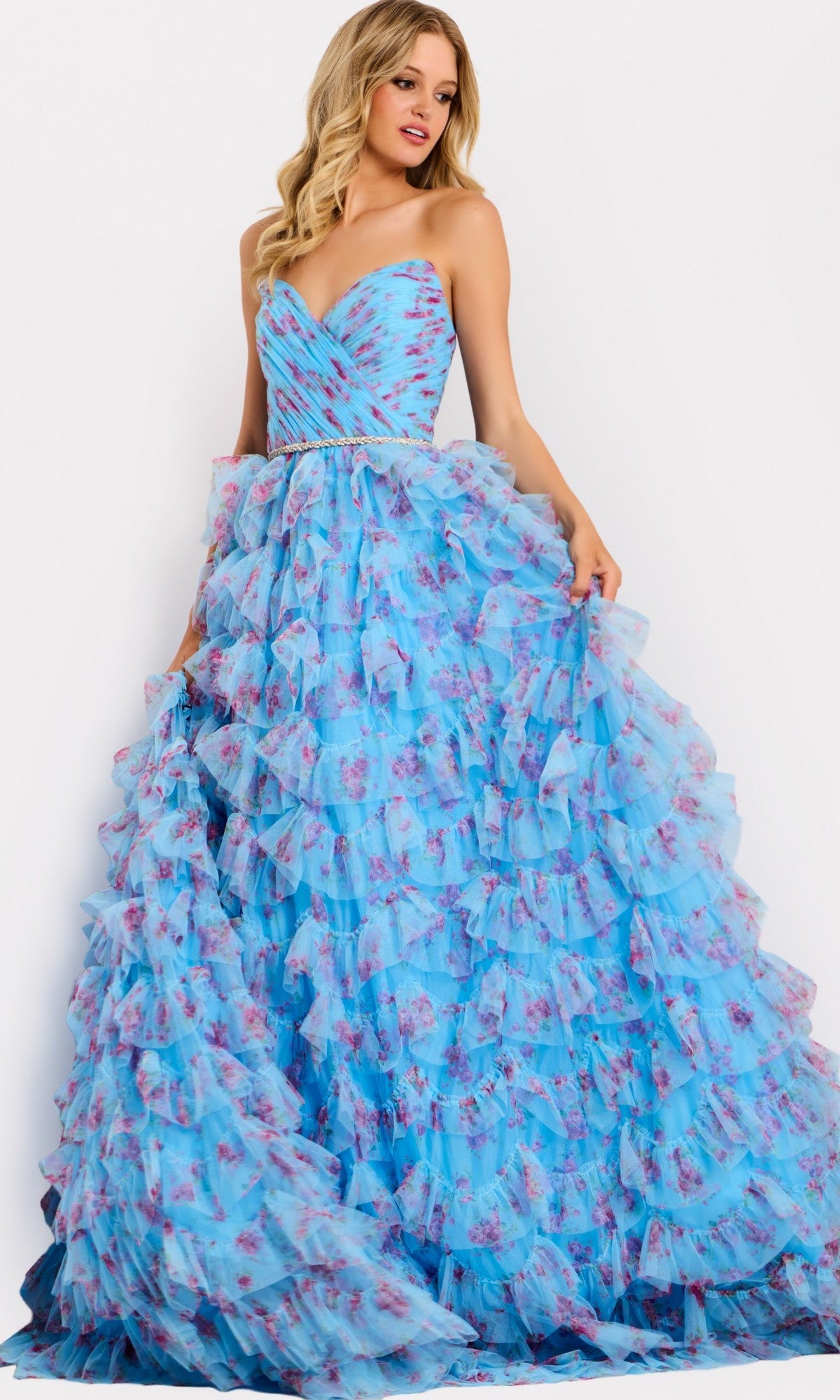 A woman models the Jovani 47267 long prom dress, a strapless ball gown with layered ruffles and a pink floral pattern, standing against a plain white background.